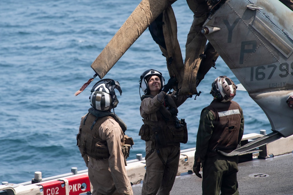Three military personnel in flight suits and helmets are working on the rotor assembly of a helicopter on the deck of a naval vessel. The helicopter, marked with 'YP' and the partial serial number '1679', appears to be a UH-1Y Venom based on visible text. The scene takes place at sea, with open water visible in the background.