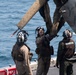 Three military personnel in flight suits and helmets are working on the rotor assembly of a helicopter on the deck of a naval vessel. The helicopter, marked with 'YP' and the partial serial number '1679', appears to be a UH-1Y Venom based on visible text. The scene takes place at sea, with open water visible in the background.