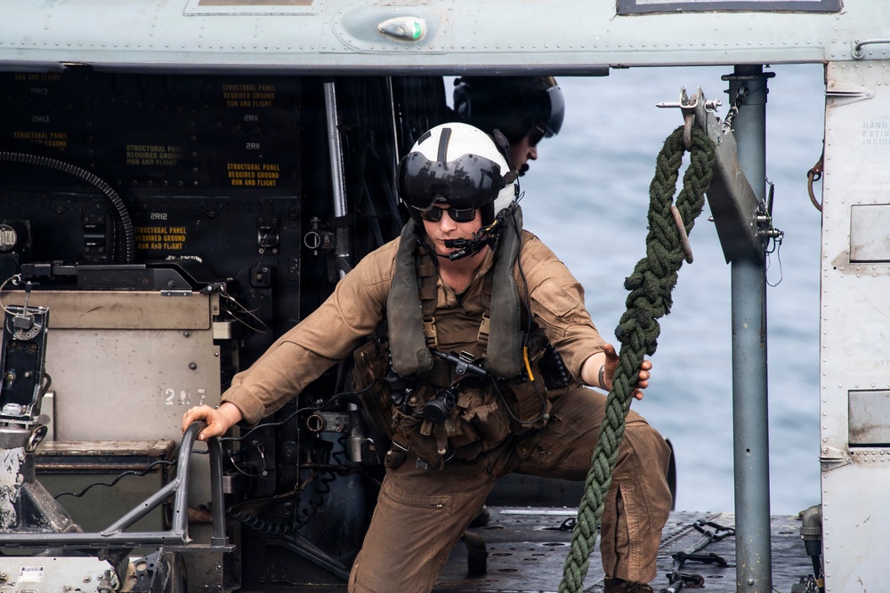 A military aviator or aircrew member in flight gear is working on the deck of a naval vessel near a helicopter. The individual is wearing a flight helmet, sunglasses, a survival vest, and tan flight suit while handling a thick rope/line. The scene appears to take place on a ship's flight deck with open ocean visible in the background.