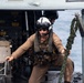 A military aviator or aircrew member in flight gear is working on the deck of a naval vessel near a helicopter. The individual is wearing a flight helmet, sunglasses, a survival vest, and tan flight suit while handling a thick rope/line. The scene appears to take place on a ship's flight deck with open ocean visible in the background.