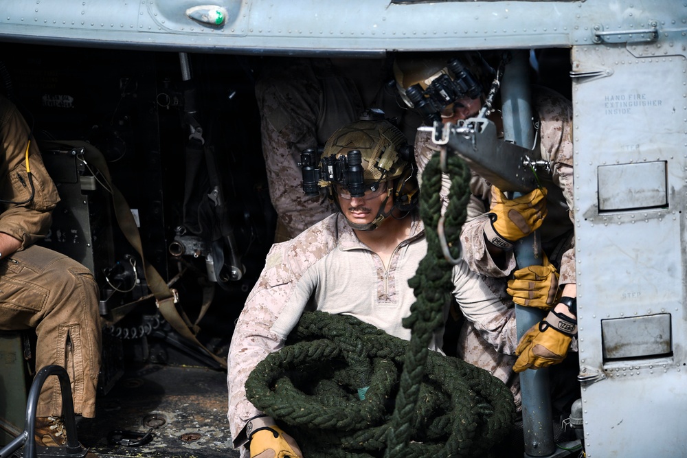 Military personnel, appearing to be U.S. Marines based on MARPAT desert camouflage uniforms, are positioned inside a helicopter doorway handling a thick coiled rope, likely for fast-rope insertion/extraction (FRIES) operations. The primary subject wears a tactical helmet mounted with night vision goggles (NVGs) and yellow tactical gloves. A second personnel member is visible behind, also equipped with NVGs and binoculars.