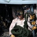 Military personnel, appearing to be U.S. Marines based on MARPAT desert camouflage uniforms, are positioned inside a helicopter doorway handling a thick coiled rope, likely for fast-rope insertion/extraction (FRIES) operations. The primary subject wears a tactical helmet mounted with night vision goggles (NVGs) and yellow tactical gloves. A second personnel member is visible behind, also equipped with NVGs and binoculars.
