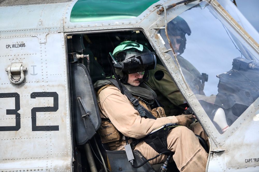 Two military aviators are seated inside a helicopter cockpit, wearing flight helmets and flight suits. The foreground pilot wears a tan/khaki flight suit with a green-visored helmet, while a second crew member is visible through the cockpit glass. Text markings on the aircraft fuselage include 'CPL WILLIAMS', 'CAPT RU[partially visible]', and the number '32'.