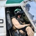 Two military aviators are seated inside a helicopter cockpit, wearing flight helmets and flight suits. The foreground pilot wears a tan/khaki flight suit with a green-visored helmet, while a second crew member is visible through the cockpit glass. Text markings on the aircraft fuselage include 'CPL WILLIAMS', 'CAPT RU[partially visible]', and the number '32'.