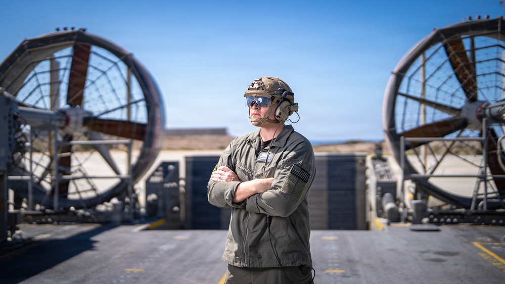 A military service member stands with arms crossed on what appears to be the deck of a Landing Craft Air Cushion (LCAC) or similar naval hovercraft vessel. The individual is wearing tactical gear including a combat helmet, communications headset, protective eyewear, and an olive drab flight/utility suit with a US flag patch. Two large propulsion fan shrouds are visible in the background against a clear blue sky.