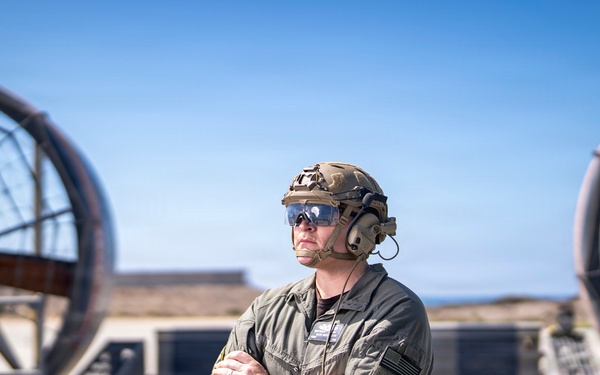 A military service member stands with arms crossed on what appears to be the deck of a Landing Craft Air Cushion (LCAC) or similar naval hovercraft vessel. The individual is wearing tactical gear including a combat helmet, communications headset, protective eyewear, and an olive drab flight/utility suit with a US flag patch. Two large propulsion fan shrouds are visible in the background against a clear blue sky.