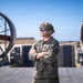 A military service member stands with arms crossed on what appears to be the deck of a Landing Craft Air Cushion (LCAC) or similar naval hovercraft vessel. The individual is wearing tactical gear including a combat helmet, communications headset, protective eyewear, and an olive drab flight/utility suit with a US flag patch. Two large propulsion fan shrouds are visible in the background against a clear blue sky.
