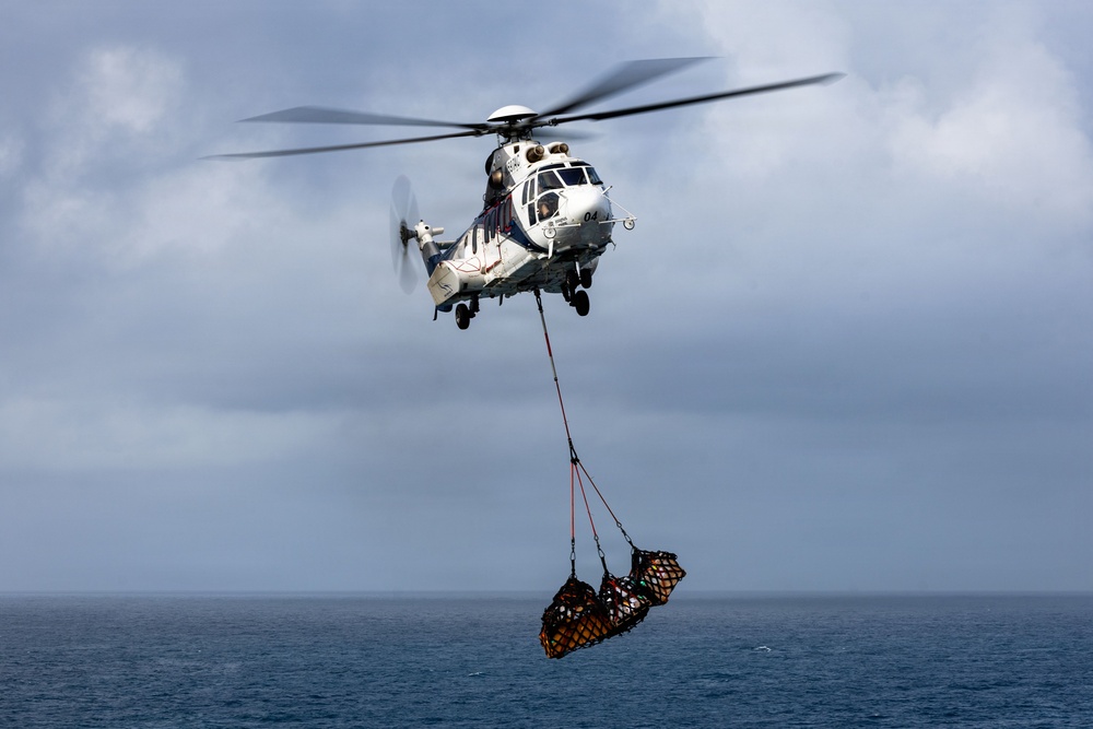 A military or coast guard helicopter is performing a vertical replenishment (VERTREP) operation over open ocean, carrying a cargo net loaded with supplies suspended beneath it. The helicopter appears to be a Super Puma or similar medium-lift rotary-wing aircraft, identifiable by its distinctive twin-engine configuration and white/blue livery with the number '04' visible. The overcast sky and open sea background suggest an at-sea logistics or resupply mission.