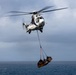 A military or coast guard helicopter is performing a vertical replenishment (VERTREP) operation over open ocean, carrying a cargo net loaded with supplies suspended beneath it. The helicopter appears to be a Super Puma or similar medium-lift rotary-wing aircraft, identifiable by its distinctive twin-engine configuration and white/blue livery with the number '04' visible. The overcast sky and open sea background suggest an at-sea logistics or resupply mission.