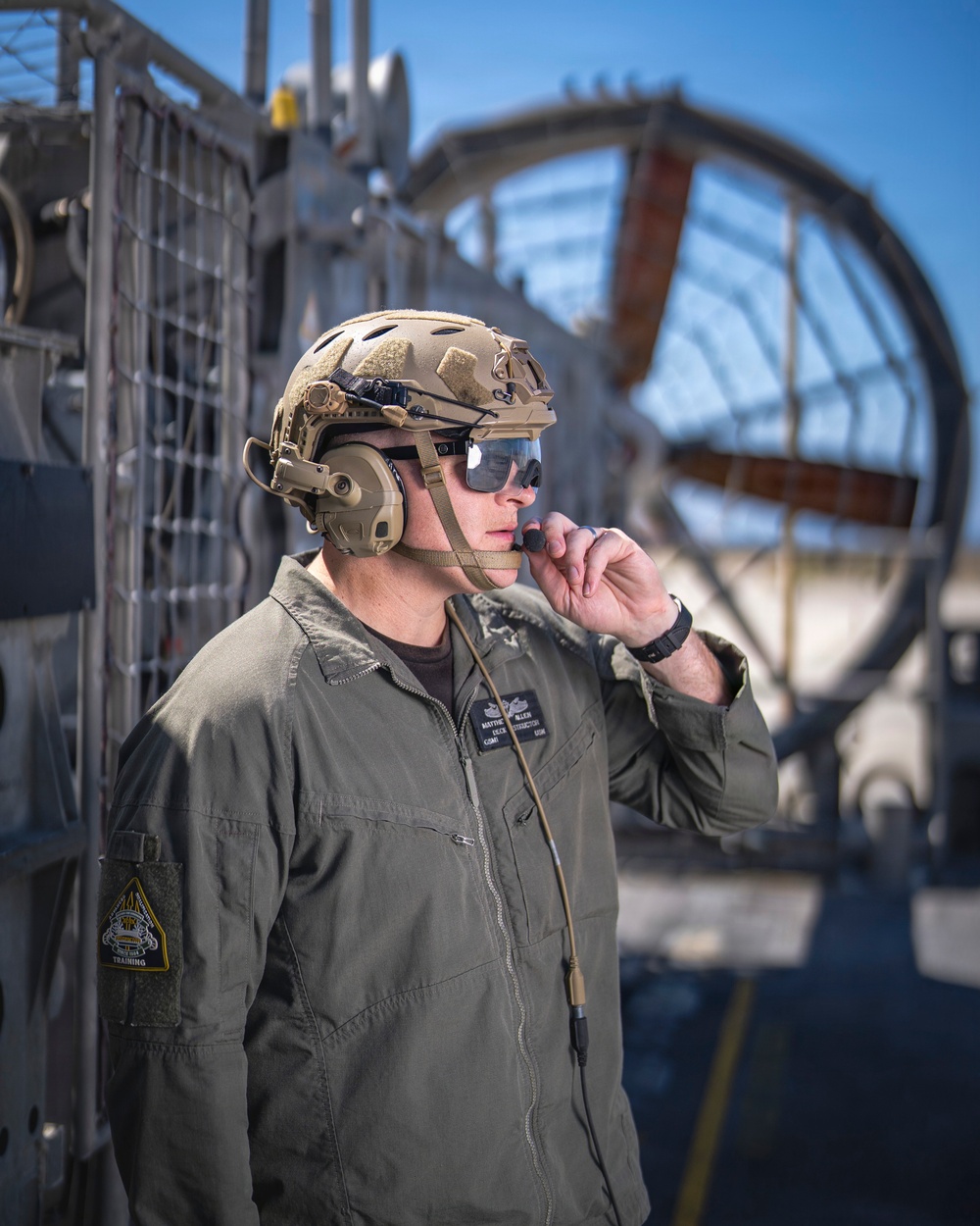 A military service member in olive drab flight suit and tan tactical helmet is shown communicating via a boom microphone/headset system. He is positioned on what appears to be a naval vessel or military platform, with large mechanical equipment visible in the background. The image is a professional, sharp portrait-style photograph taken in daylight.