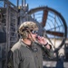 A military service member in olive drab flight suit and tan tactical helmet is shown communicating via a boom microphone/headset system. He is positioned on what appears to be a naval vessel or military platform, with large mechanical equipment visible in the background. The image is a professional, sharp portrait-style photograph taken in daylight.