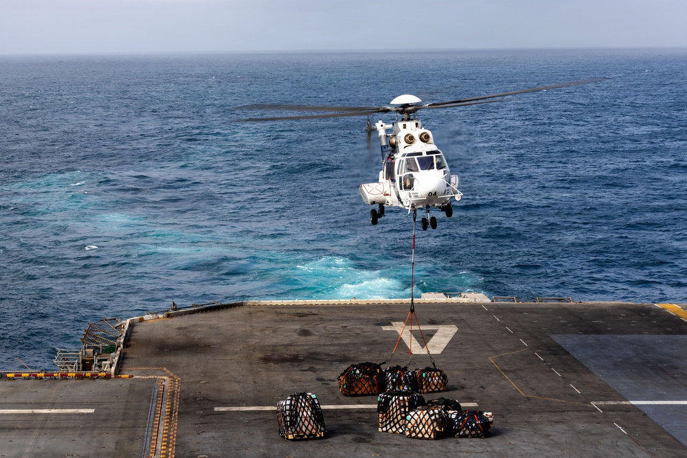 A white military or coast guard helicopter is hovering over the flight deck of a naval vessel at sea, performing a vertical replenishment (VERTREP) operation. The helicopter, marked with the number '04', is connected via a sling line to multiple cargo nets filled with supplies on the deck below. The operation is taking place in open ocean conditions with calm to moderate seas visible in the background.