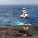 A white military or coast guard helicopter is hovering over the flight deck of a naval vessel at sea, performing a vertical replenishment (VERTREP) operation. The helicopter, marked with the number '04', is connected via a sling line to multiple cargo nets filled with supplies on the deck below. The operation is taking place in open ocean conditions with calm to moderate seas visible in the background.