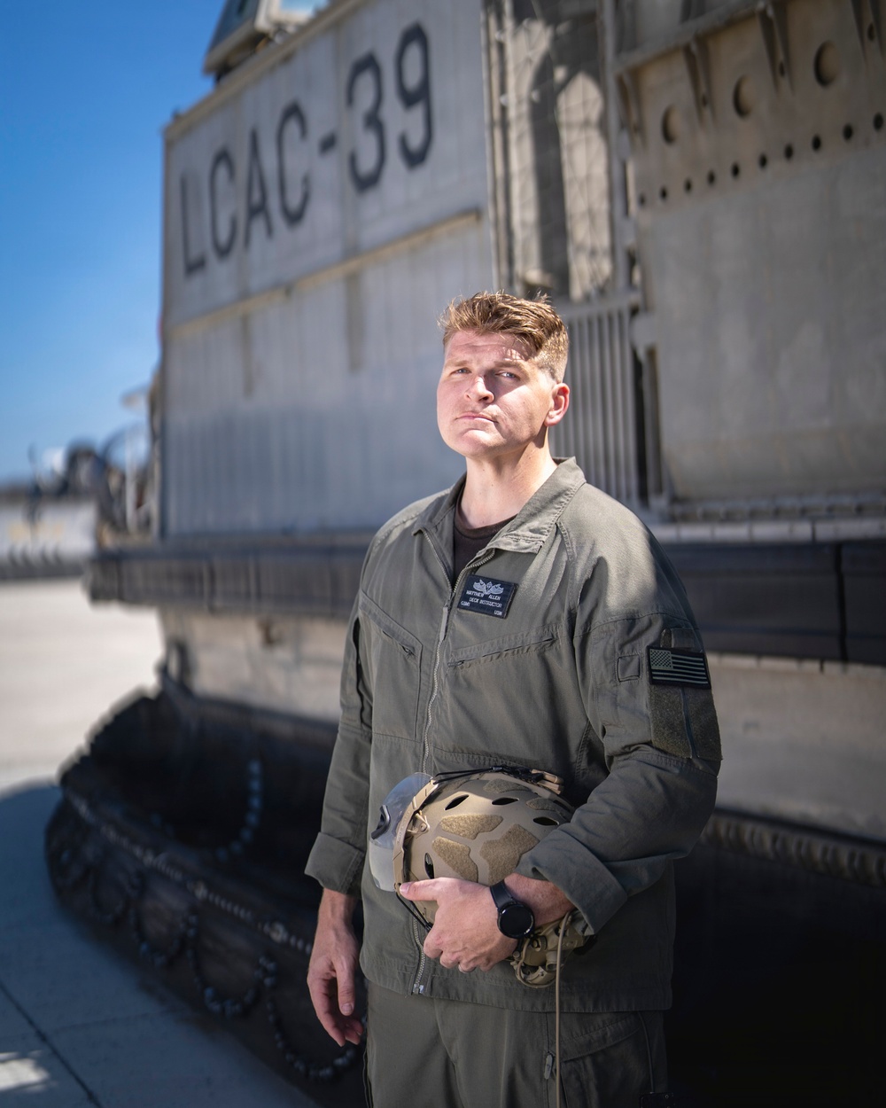 A military service member in olive drab coveralls stands in front of Landing Craft Air Cushion number 39 (LCAC-39), a U.S. Navy hovercraft. He is holding a tactical helmet and wearing a name tag with qualification wings on his chest, along with a U.S. flag patch on his right sleeve. The image appears to be an official or professional portrait-style photograph taken outdoors in clear daylight.