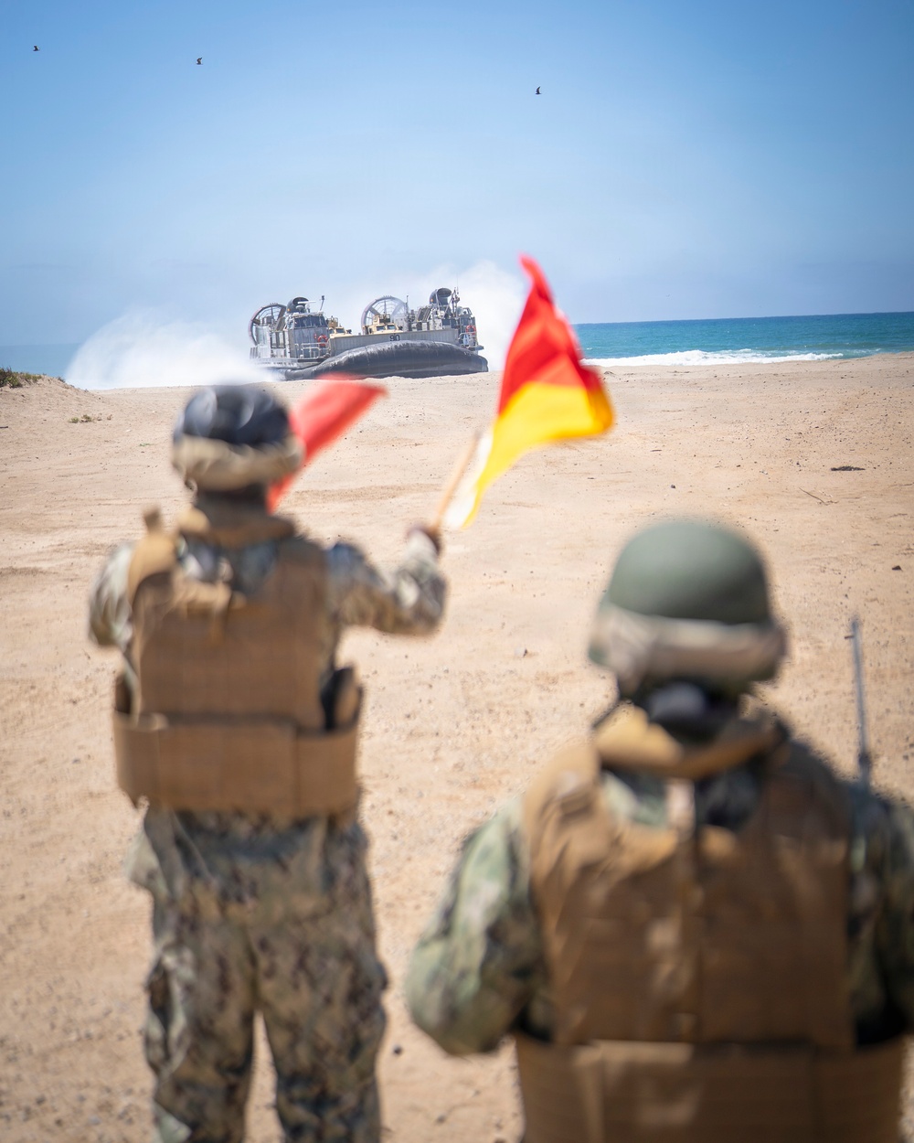 Two military personnel in combat gear stand on a sandy beach, using signal flags to guide a Landing Craft Air Cushion (LCAC) hovercraft coming ashore from the ocean. The scene depicts a amphibious landing operation, with one service member actively waving red and yellow signal flags toward the approaching vessel. The foreground subjects are slightly out of focus while the LCAC in the background is sharply rendered.