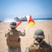 Two military personnel in combat gear stand on a sandy beach, using signal flags to guide a Landing Craft Air Cushion (LCAC) hovercraft coming ashore from the ocean. The scene depicts a amphibious landing operation, with one service member actively waving red and yellow signal flags toward the approaching vessel. The foreground subjects are slightly out of focus while the LCAC in the background is sharply rendered.