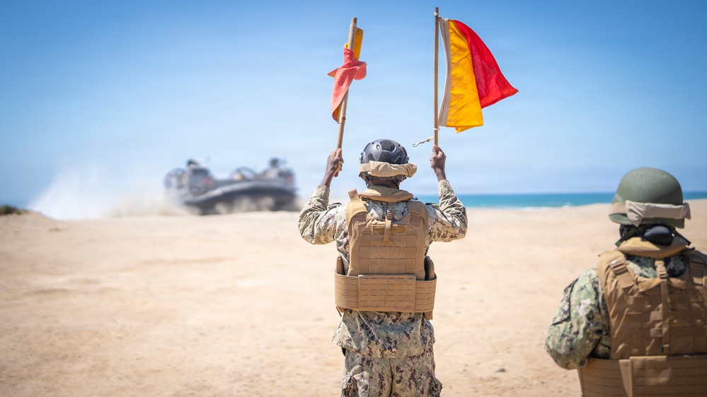 Military personnel on a sandy beach are using semaphore flags to guide a Landing Craft Air Cushion (LCAC) hovercraft onto shore. The central figure, wearing desert camouflage and a tactical vest, holds two flags — one red and one red-and-yellow — raised above their head. A second service member in camouflage and a green helmet observes from the right foreground.