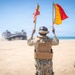Military personnel on a sandy beach are using semaphore flags to guide a Landing Craft Air Cushion (LCAC) hovercraft onto shore. The central figure, wearing desert camouflage and a tactical vest, holds two flags — one red and one red-and-yellow — raised above their head. A second service member in camouflage and a green helmet observes from the right foreground.