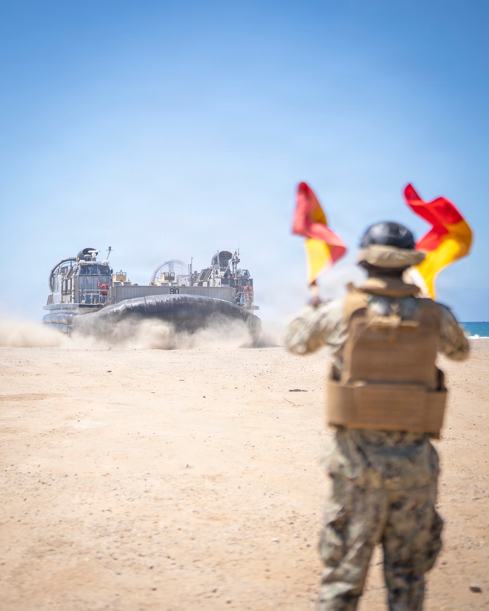 A military personnel in camouflage uniform and body armor is using semaphore flags (red and yellow) to guide a Landing Craft Air Cushion (LCAC) hovercraft onto a sandy beach. The LCAC, marked with the number '91', is approaching from the water, kicking up dust and spray as it travels across the beach. This appears to be an amphibious landing operation or military exercise.