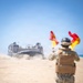 A military personnel in camouflage uniform and body armor is using semaphore flags (red and yellow) to guide a Landing Craft Air Cushion (LCAC) hovercraft onto a sandy beach. The LCAC, marked with the number '91', is approaching from the water, kicking up dust and spray as it travels across the beach. This appears to be an amphibious landing operation or military exercise.