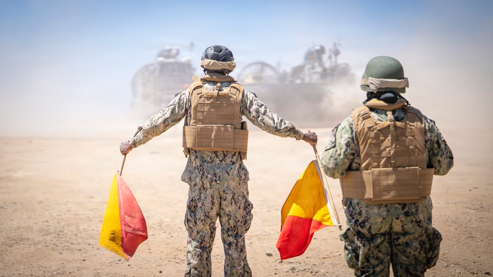 Two military personnel in camouflage uniforms and tactical vests are using semaphore-style signal flags (red and yellow) to guide a large armored vehicle in a dusty desert environment. The scene appears to be a military exercise or operational movement, with significant dust kicked up by the approaching vehicle. Both individuals are viewed from behind, arms extended holding the signal flags.