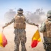 Two military personnel in camouflage uniforms and tactical vests are using semaphore-style signal flags (red and yellow) to guide a large armored vehicle in a dusty desert environment. The scene appears to be a military exercise or operational movement, with significant dust kicked up by the approaching vehicle. Both individuals are viewed from behind, arms extended holding the signal flags.