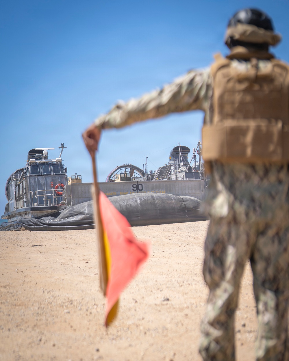 A military service member in camouflage uniform and tactical vest stands on a sandy beach, holding a signal flag while directing a Landing Craft Air Cushion (LCAC) hovercraft numbered '90' toward the shore. The scene depicts an amphibious landing operation with the LCAC beached or approaching the shoreline. The foreground subject is intentionally blurred with the LCAC in sharp focus in the background.