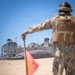 A military service member in camouflage uniform and tactical vest stands on a sandy beach, holding a signal flag while directing a Landing Craft Air Cushion (LCAC) hovercraft numbered '90' toward the shore. The scene depicts an amphibious landing operation with the LCAC beached or approaching the shoreline. The foreground subject is intentionally blurred with the LCAC in sharp focus in the background.