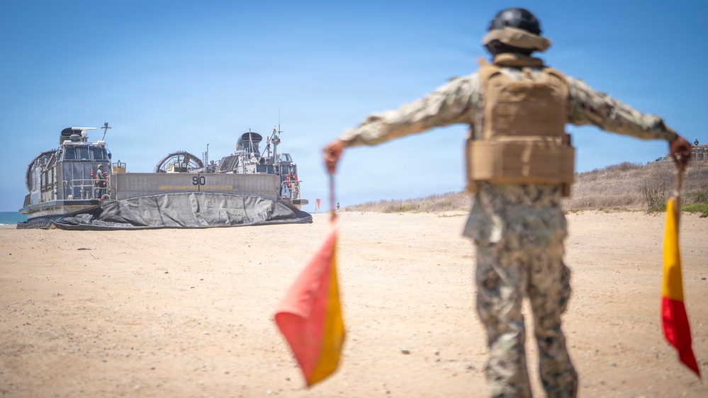 A military service member in camouflage uniform and tactical vest stands on a sandy beach, using signal flags to guide a Landing Craft Air Cushion (LCAC) hovercraft numbered '90' onto the shore. The scene depicts a standard amphibious landing operation with the LCAC approaching from the water's edge. The setting appears to be a training or operational beach landing exercise under clear skies.