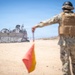 A military service member in camouflage uniform and tactical vest stands on a sandy beach, using signal flags to guide a Landing Craft Air Cushion (LCAC) hovercraft numbered '90' onto the shore. The scene depicts a standard amphibious landing operation with the LCAC approaching from the water's edge. The setting appears to be a training or operational beach landing exercise under clear skies.