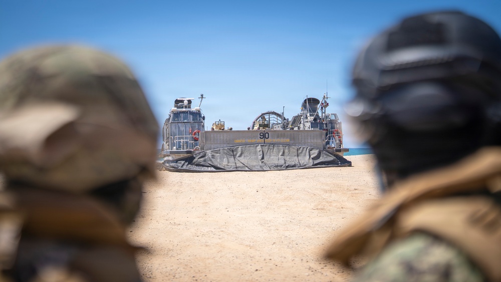 Two military personnel in helmets and camouflage gear observe a Landing Craft Air Cushion (LCAC) numbered '90' that has beached on a sandy shore. The hovercraft, with its deflated skirt visible, appears to have completed or is preparing for an amphibious landing operation. The scene takes place under clear blue skies in an arid coastal environment.