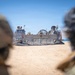 Two military personnel in helmets and camouflage gear observe a Landing Craft Air Cushion (LCAC) numbered '90' that has beached on a sandy shore. The hovercraft, with its deflated skirt visible, appears to have completed or is preparing for an amphibious landing operation. The scene takes place under clear blue skies in an arid coastal environment.