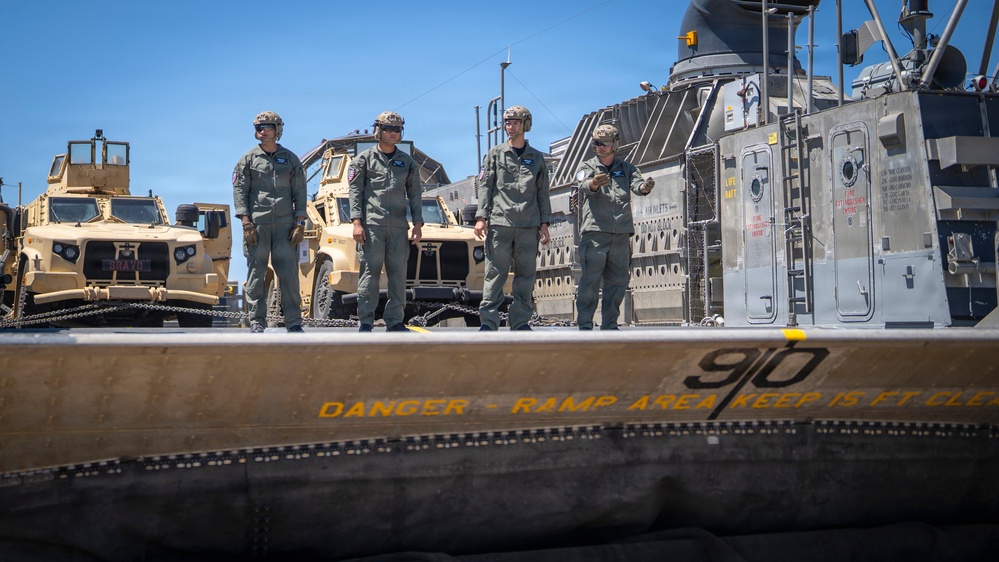 Four military personnel in olive/sage green flight suits and helmets stand on what appears to be a military vessel or landing craft ramp area. Behind them are several tan/desert-colored military tactical vehicles, likely Joint Light Tactical Vehicles (JLTVs) or MRAPs, secured with chains. The foreground shows a safety warning stenciled in yellow reading 'DANGER - RAMP AREA KEEP 15 FT CLE[AR]' along with the number '9|0'.