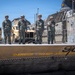Four military personnel in olive/sage green flight suits and helmets stand on what appears to be a military vessel or landing craft ramp area. Behind them are several tan/desert-colored military tactical vehicles, likely Joint Light Tactical Vehicles (JLTVs) or MRAPs, secured with chains. The foreground shows a safety warning stenciled in yellow reading 'DANGER - RAMP AREA KEEP 15 FT CLE[AR]' along with the number '9|0'.
