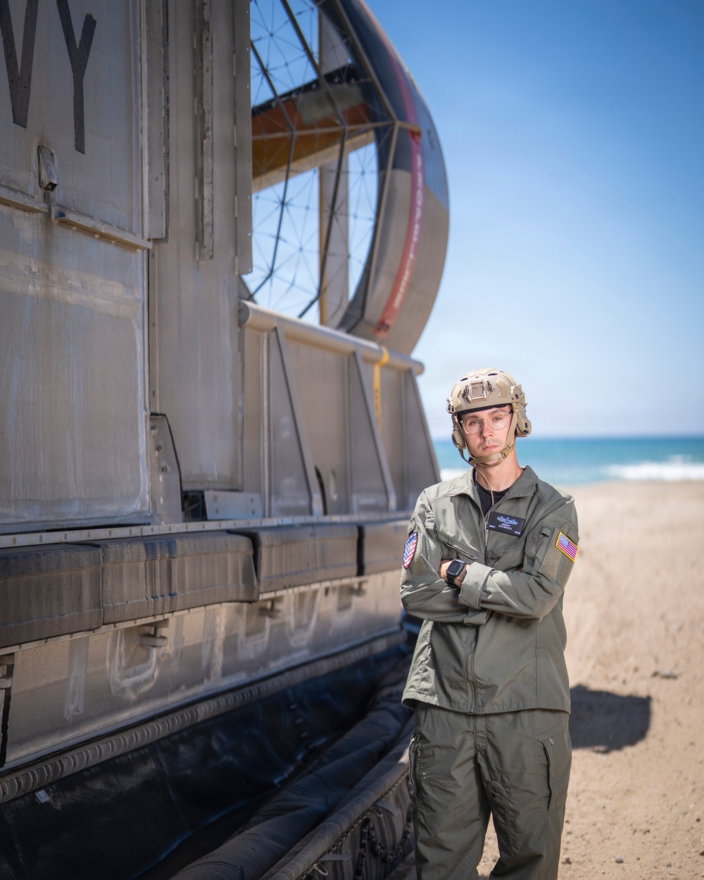 A military service member in olive green flight suit stands with arms crossed beside a large naval vessel on a sandy beach. The vessel displays partial lettering 'VY' consistent with a U.S. Navy LCAC (Landing Craft Air Cushion) hovercraft, identifiable by its inflatable skirt and large fan shroud. The setting appears to be a coastal amphibious operations environment under clear blue skies.
