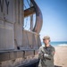 A military service member in olive green flight suit stands with arms crossed beside a large naval vessel on a sandy beach. The vessel displays partial lettering 'VY' consistent with a U.S. Navy LCAC (Landing Craft Air Cushion) hovercraft, identifiable by its inflatable skirt and large fan shroud. The setting appears to be a coastal amphibious operations environment under clear blue skies.