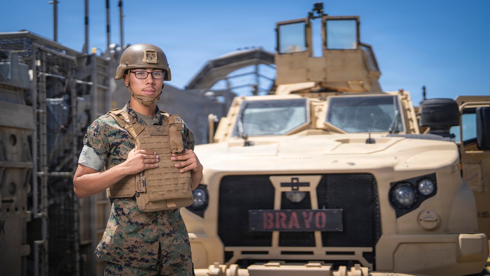 A U.S. Marine in full combat gear stands in front of a tan/desert-colored military vehicle labeled 'BRAVO'. The service member is wearing MARPAT digital camouflage, a tactical vest, and a combat helmet with glasses. The setting appears to be a military staging or motor pool area under clear blue skies.
