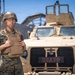 A U.S. Marine in full combat gear stands in front of a tan/desert-colored military vehicle labeled 'BRAVO'. The service member is wearing MARPAT digital camouflage, a tactical vest, and a combat helmet with glasses. The setting appears to be a military staging or motor pool area under clear blue skies.