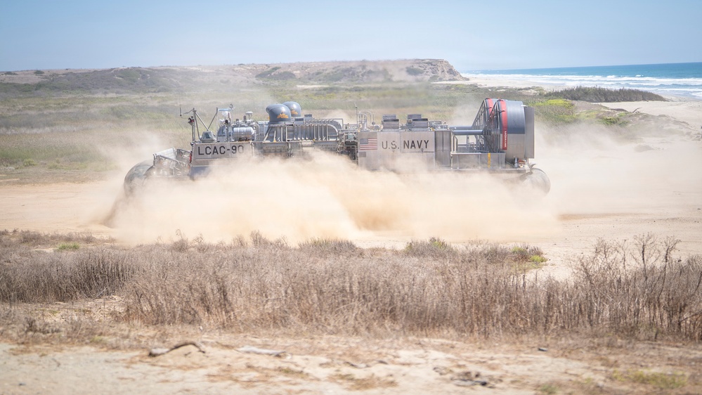A U.S. Navy Landing Craft Air Cushion (LCAC-90) hovercraft is operating on a sandy beach terrain, kicking up large dust clouds as it moves inland from the ocean. The vessel is clearly marked with 'LCAC-90' and 'U.S. NAVY' on its hull, along with an American flag decal. The coastal environment features sandy dunes, dry scrub vegetation, and the Pacific Ocean visible in the background.