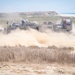 A U.S. Navy Landing Craft Air Cushion (LCAC-90) hovercraft is operating on a sandy beach terrain, kicking up large dust clouds as it moves inland from the ocean. The vessel is clearly marked with 'LCAC-90' and 'U.S. NAVY' on its hull, along with an American flag decal. The coastal environment features sandy dunes, dry scrub vegetation, and the Pacific Ocean visible in the background.
