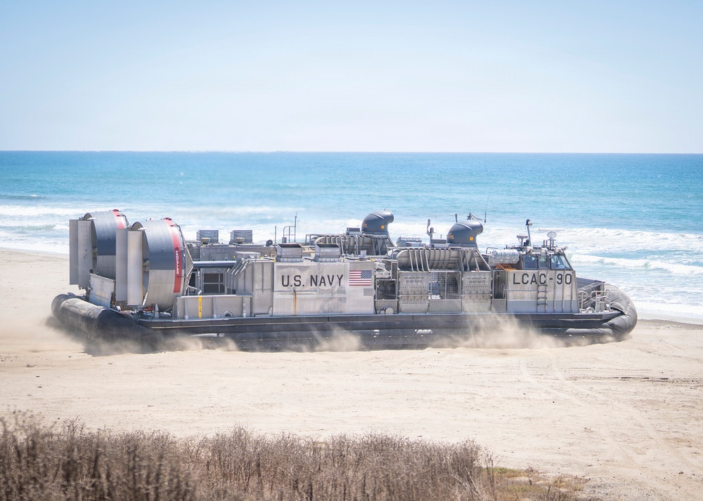 A U.S. Navy Landing Craft Air Cushion (LCAC-90) hovercraft is operating on a sandy beach, transitioning between land and sea. The vessel is visibly hovering above the sand, generating dust clouds beneath its skirt. This appears to be a military amphibious operation or training exercise.