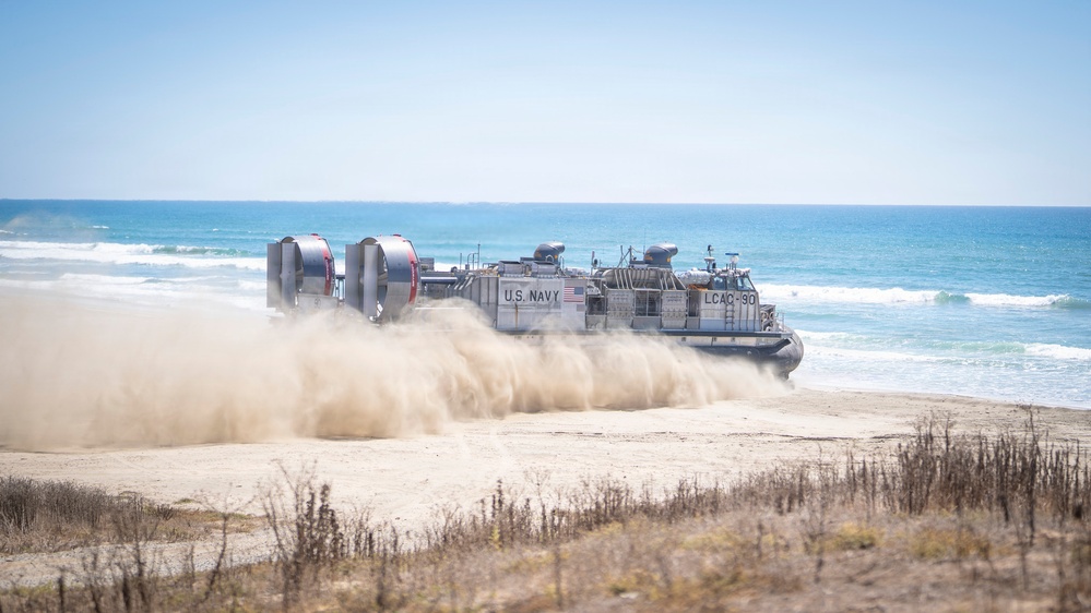 A U.S. Navy Landing Craft Air Cushion (LCAC-90) is shown operating on a sandy beach, kicking up large clouds of dust and sand as it moves across the shoreline. The hovercraft is transitioning between the ocean and the beach, demonstrating its amphibious capability. The ocean with breaking waves is visible in the background under a clear blue sky.