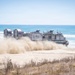 A U.S. Navy Landing Craft Air Cushion (LCAC-90) is shown operating on a sandy beach, kicking up large clouds of dust and sand as it moves across the shoreline. The hovercraft is transitioning between the ocean and the beach, demonstrating its amphibious capability. The ocean with breaking waves is visible in the background under a clear blue sky.