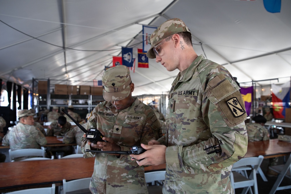 Two U.S. Army soldiers in OCP (Operational Camouflage Pattern) uniforms are examining what appears to be a drone controller or radio communication device inside a large military tent. The foreground soldier wears a 1st Cavalry Division patch on his right sleeve and safety glasses, while both soldiers appear engaged in equipment operation or training. The setting appears to be a field operations center or command post with multiple personnel and military unit flags visible in the background.