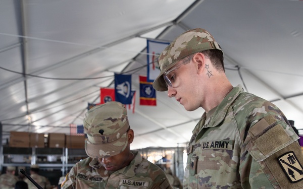 Two U.S. Army soldiers in OCP (Operational Camouflage Pattern) uniforms are examining what appears to be a drone controller or radio communication device inside a large military tent. The foreground soldier wears a 1st Cavalry Division patch on his right sleeve and safety glasses, while both soldiers appear engaged in equipment operation or training. The setting appears to be a field operations center or command post with multiple personnel and military unit flags visible in the background.