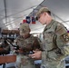 Two U.S. Army soldiers in OCP (Operational Camouflage Pattern) uniforms are examining what appears to be a drone controller or radio communication device inside a large military tent. The foreground soldier wears a 1st Cavalry Division patch on his right sleeve and safety glasses, while both soldiers appear engaged in equipment operation or training. The setting appears to be a field operations center or command post with multiple personnel and military unit flags visible in the background.