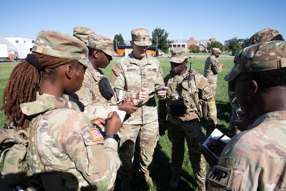 A group of U.S. Army soldiers in OCP (Operational Camouflage Pattern) uniforms are gathered outdoors on a grassy field in what appears to be a briefing or instruction session. A central soldier, identified by nametape as 'NOONAN' and wearing a 'U.S. ARMY' chest tape, appears to be leading the discussion while others take notes or observe. The setting appears to be a military installation or training area.
