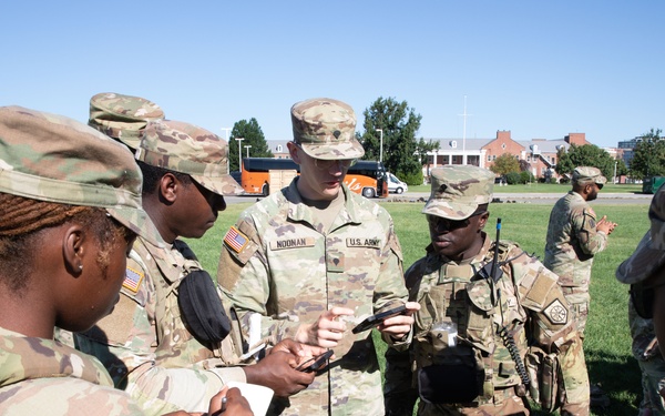 A group of U.S. Army soldiers in OCP (Operational Camouflage Pattern) uniforms are gathered outdoors on a grassy field in what appears to be a briefing or instruction session. A central soldier, identified by nametape as 'NOONAN' and wearing a 'U.S. ARMY' chest tape, appears to be leading the discussion while others take notes or observe. The setting appears to be a military installation or training area.