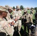 A group of U.S. Army soldiers in OCP (Operational Camouflage Pattern) uniforms are gathered outdoors on a grassy field in what appears to be a briefing or instruction session. A central soldier, identified by nametape as 'NOONAN' and wearing a 'U.S. ARMY' chest tape, appears to be leading the discussion while others take notes or observe. The setting appears to be a military installation or training area.