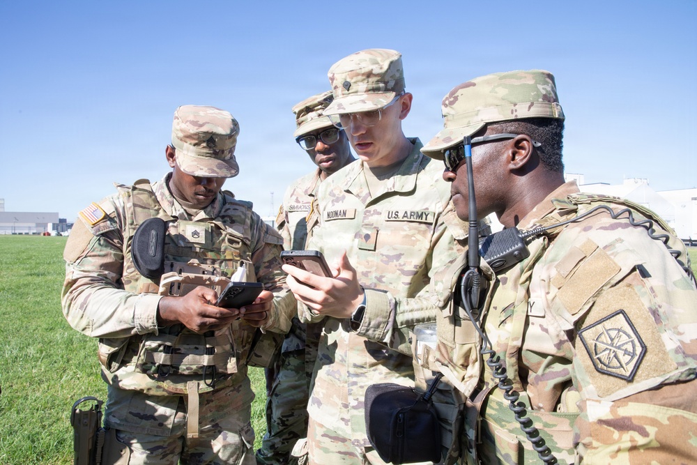 Four U.S. Army soldiers in OCP (Operational Camouflage Pattern) uniforms are gathered outdoors, reviewing information on mobile devices. The soldiers appear to be coordinating or sharing information, with at least two holding smartphones and one equipped with a radio. The setting appears to be a military installation or training area with a clear blue sky and green grass visible in the background.